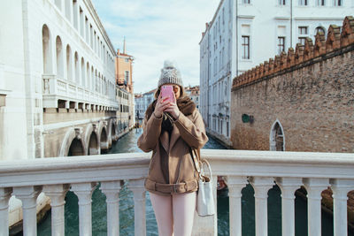 Young woman using phone while standing by canal in city