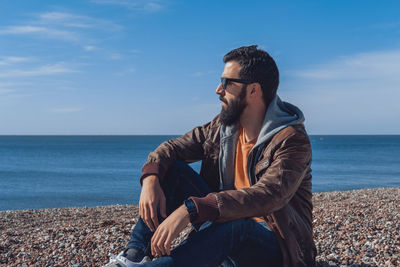 Young man sitting on shore at beach against sky