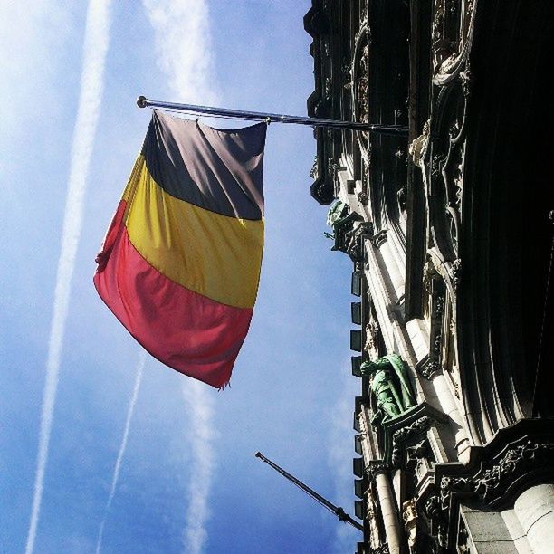 low angle view, building exterior, architecture, built structure, sky, flag, hanging, blue, outdoors, day, no people, clear sky, identity, patriotism, pole, building, national flag, rope, city, sunlight