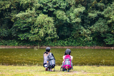 Rear view of women crouching on grass by lake against trees