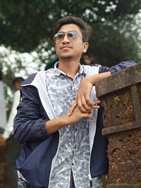 Young man wearing sunglasses standing by wall against trees