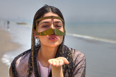 Portrait of young woman wearing traditional clothing blowing kiss at beach against sky