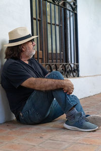 Man in a black shirt, hat and slippers sitting in front of a house