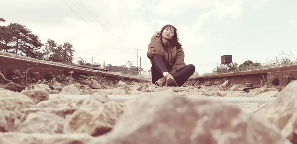 Young woman sitting on railroad track against sky