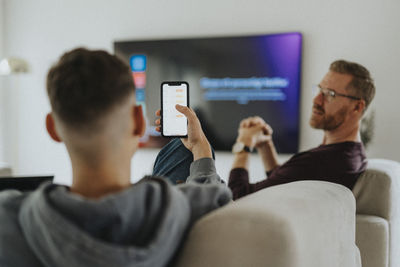 Teenage boy with mobile phone talking to father while sitting in living room