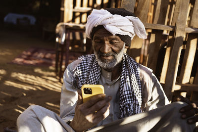 Elderly nubian male in traditional clothes and turban sitting in yard and using mobile phone on sunny day