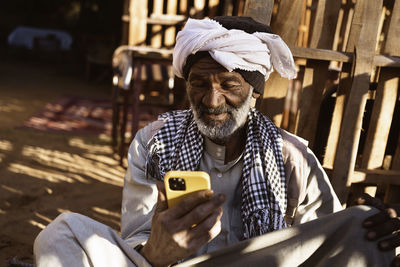 Elderly nubian male in traditional clothes and turban sitting in yard and using mobile phone on sunny day