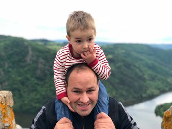 Portrait of smiling man carrying son on shoulders against mountains