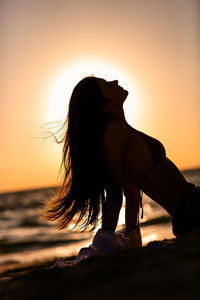 Side view of woman sitting on beach against sky during sunset