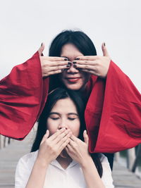 Playful female friends covering eyes and mouth with hands against clear sky