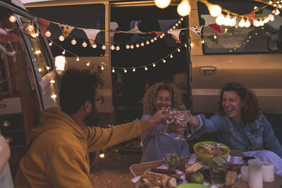 Male and female friends toasting while sitting by motor home during sunset