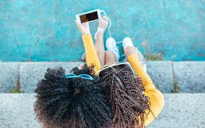 From above of unrecognizable african american female with curly hair sitting on stone stairs with woman with braids and watching video together on cellphone