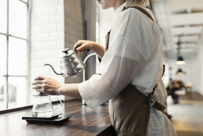 Side view midsection of female barista pouring boiling water in coffee filter at cafe counter