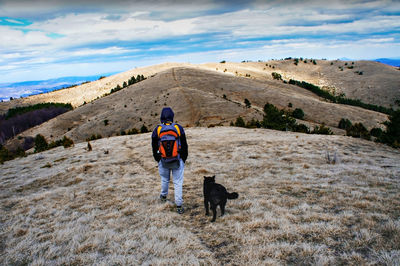 Young man from behind with backpack walking in hills with black dog against blue cloudy sky. hiking