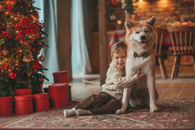 Candid authentic happy little boy in knitted beige sweater hugs dog with bow tie at home on xmas