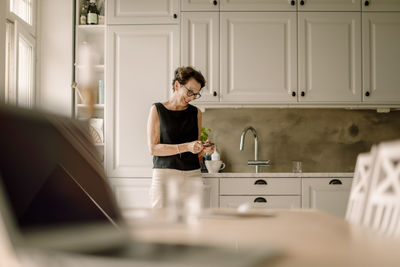 Retired senior woman using smart phone while leaning on kitchen counter