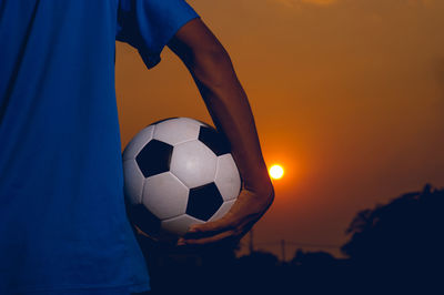 Cropped hand holding soccer ball against sky during sunset