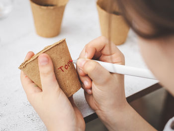 The hands of a caucasian girl hold a cardboard glass and sign the word tomato with a red marker