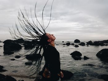 Side view of woman tossing dreadlocks at beach against sky