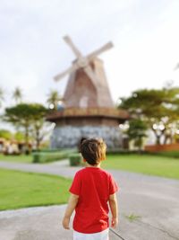 Rear view of boy looking at traditional windmill while standing on footpath against sky