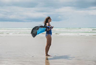 Side view of woman in swimwear holding sarong while standing at beach against cloudy sky during sunny day