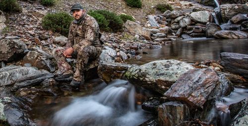 Full length of thoughtful man sitting on rock by stream in forest