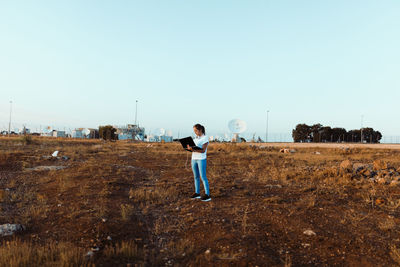 Full length of woman using laptop while standing on field against clear sky