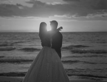 Married couple standing on beach against sky during sunset