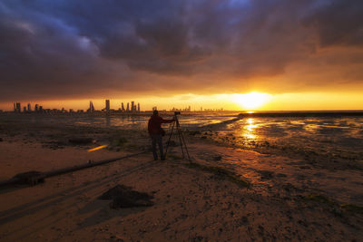 Rear view of man standing with camera at beach against cloudy sky during sunset