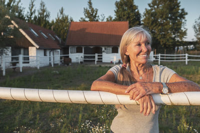 Smiling senior female farm worker leaning on fence at farm