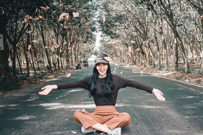 Portrait of smiling young woman throwing leaves while sitting on road amidst trees in forest