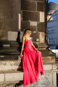Full length of young woman in pink evening gown sitting against columns