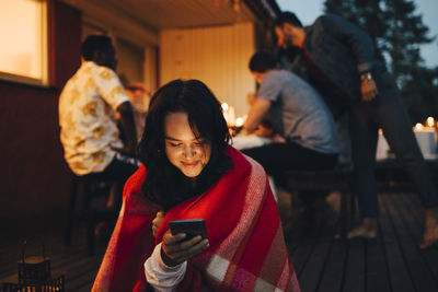 Smiling young woman using mobile phone while friends in background during dinner party