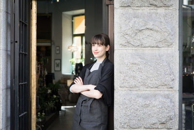 Smiling confident young female entrepreneur standing with arms crossed at store doorway