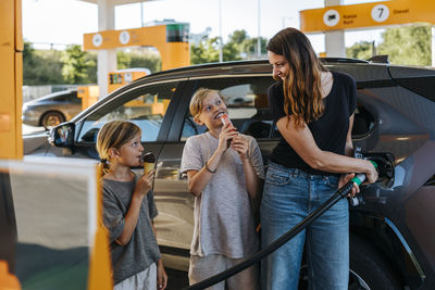 Smiling woman refueling car and standing with sons eating snacks at gas station