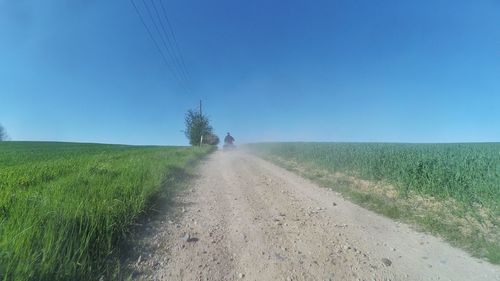 Rear view of man driving quadbike on dirt road amidst grassy field against clear blue sky