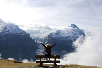 Rear view of woman sitting on bench against snow covered mountains and sky