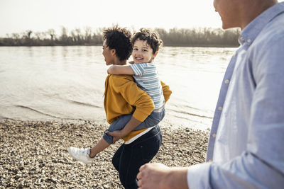 Cheerful boy enjoying piggybacked on mother walking by father at lakeside