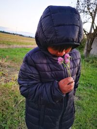 Cute boy wearing padded jacket smelling flower on field against sky