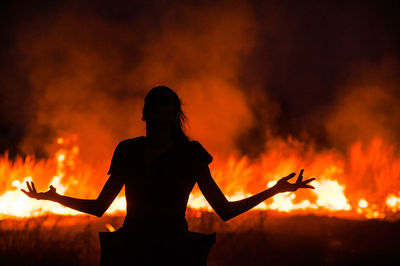Rear view of silhouette woman with arms outstretched in front of fire on field