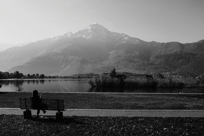 Rear view of man sitting on bench by lake against sky