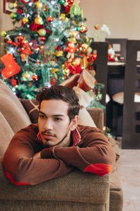 Thoughtful man lying on sofa against decorated christmas tree at home