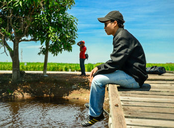 Man sitting on pier over lake while friend photographing in background