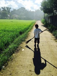 Rear view of boy holding broom while walking on footpath against sky