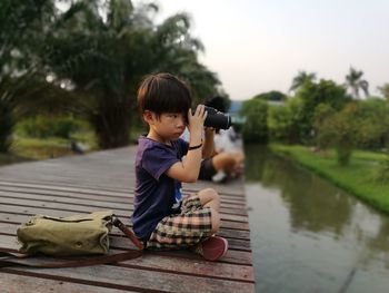 Boy photographing while sitting on pier over lake