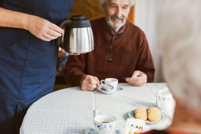 Midsection of caretaker holding coffee pot while senior couple playing cards in nursing home