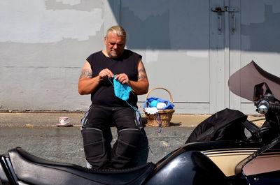 Man knitting wool while sitting against wall