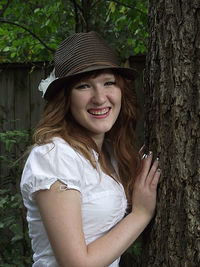 Young woman wearing hat standing by tree trunk in park