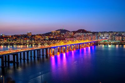 Illuminated buildings by river against blue sky at night