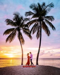 Romantic couple standing at beach against sky during sunset